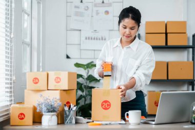 Beyaz gömlekli genç bir kadın, karton kutular paketliyor. Ofis ortamında taşınmak için. Masasında dizüstü bilgisayar ve ofis malzemeleri var..