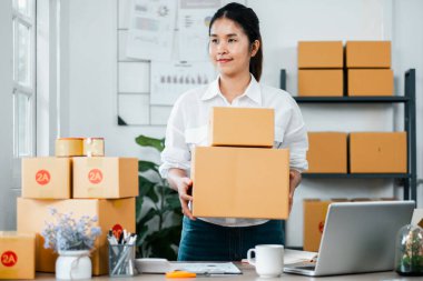 Genç bir kadın işyeri sahibi, kutularla ve ofis malzemeleriyle çevrili modern ev ofisinde nakliye için paketler düzenler ve hazırlar..
