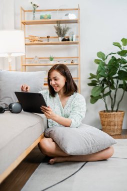 Yerde oturan genç bir kadın, kanepeye yaslanmış, rahat, modern bir oturma odasında tablet kullanıyor, iç mekan bitkileri ve şık dekorları olan..