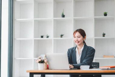 Modern bir ofiste dizüstü bilgisayarıyla çalışan genç, profesyonel bir kadın. Minimalist dekorasyonu ve doğal ışığı var. Bitkilerle çevrili..