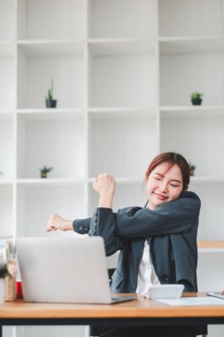 Genç profesyonel bir kadın, raflarında minimalist dekor ve küçük yeşil bitkilerle modern bir ofiste masasına uzanır..