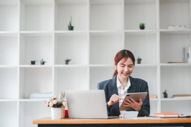 Modern ofis ortamında, bilgisayarında tablet kullanan ve arka planda raflar düzenleyen genç profesyonel bir kadın..