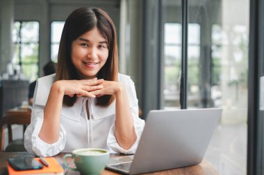 Modern bir kafede dizüstü bilgisayarda çalışırken gülümseyen genç bir kadın, bir fincan kahvenin tadını çıkarıyor..