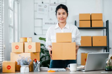 Ev ofisinde karton kutu taşıyan genç bir kadın, online bir iş ve e-ticaret ortamını temsil ediyor..