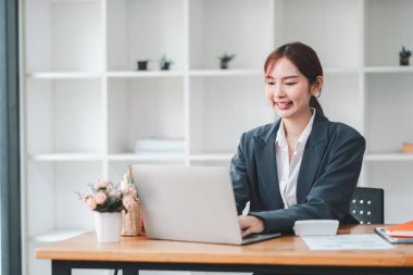 Çağdaş ofis ortamında dizüstü bilgisayarla çalışan iş elbiseli gülümseyen genç bir kadın..