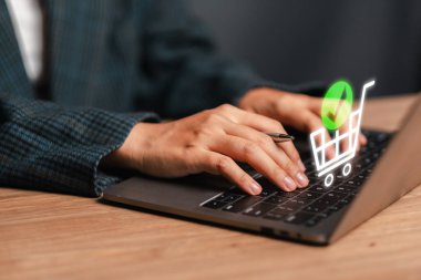 Çevrimiçi alışveriş ve e-ticareti temsil eden dijital alışveriş arabası ikonu olan bir dizüstü bilgisayarda yazı yazan ellerin yakın çekimi.