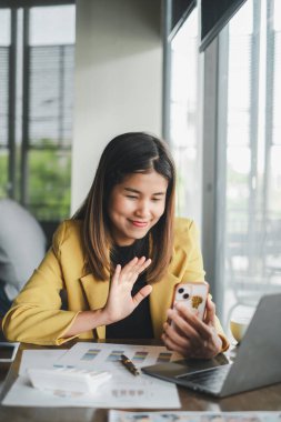 Sarı spor ceketli genç bir kadın modern ofis ortamında çalışırken akıllı telefonundan arıyor..