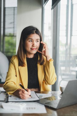 Sarı ceketli genç profesyonel bir kadın telefonla konuşuyor ve modern bir ofis masasında dizüstü bilgisayar ve belgelerle notlar alıyor..