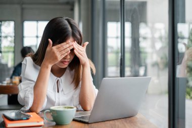Beyaz bluzlu genç bir kadın modern bir kafede dizüstü bilgisayarı üzerinde çalışırken stresli görünüyor. Masasında kahve fincanı var..