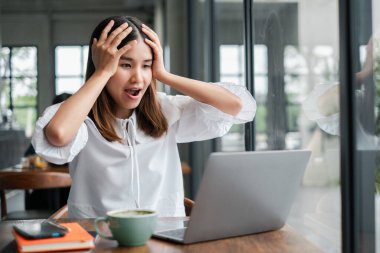 Beyaz bluzlu genç bir kadın modern bir kafede dizüstü bilgisayarına bakarken şaşırır. Masanın üzerinde bir fincan kahve ve bir defter var..