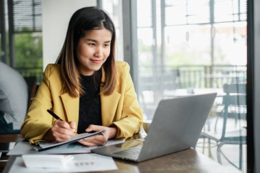 Sarı ceketli genç bir kadın dizüstü bilgisayarda çalışıyor ve geniş pencereli modern ofis ortamında notlar alıyor..