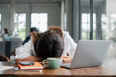 Masasında kafası olan yorgun bir kadın, dizüstü bilgisayar, kahve ve defterle çevrili, parlak, modern bir ofis ortamında..