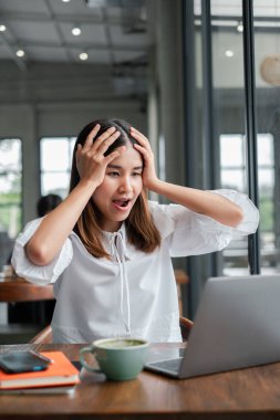 Beyaz bluzlu genç bir kadın, modern bir kafe ortamında dizüstü bilgisayarına bakarken şok oluyor ve şaşırıyor..