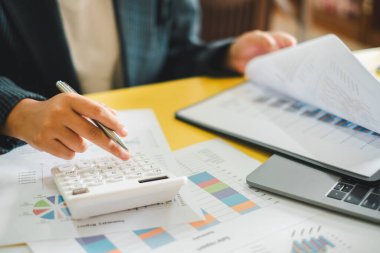 Bir iş adamı hesap makinesi kullanır ve finansal belgeleri bir masada inceler, veri analizi ve finansal planlamaya odaklanır..