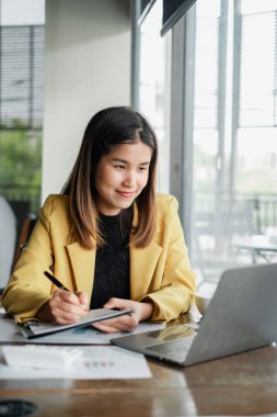 Sarı ceketli genç bir kadın büyük pencereleri ve doğal ışığı olan parlak, modern bir ofis alanında dizüstü bilgisayar üzerinde çalışıyor..
