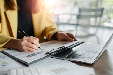 Sarı ceketli bir iş adamı dizüstü bilgisayardaki veri ve çizelgeleri ve modern ofis ortamındaki belgeleri analiz ediyor..