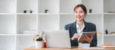Modern bir ofis ortamında çalışırken tablet kullanan gülümseyen profesyonel bir kadın ve arka planda dizüstü bilgisayarlar..