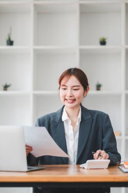 Masasında dizüstü bilgisayarı ve modern ofis ortamında dokümanlarıyla çalışan spor ceketli genç bir kadın..