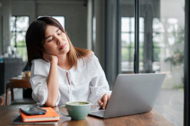 Beyaz bluzlu genç bir kadın rahat bir kafede dizüstü bilgisayarda çalışıyor, kahve molası veriyor ve büyük bir pencerede dinleniyor..