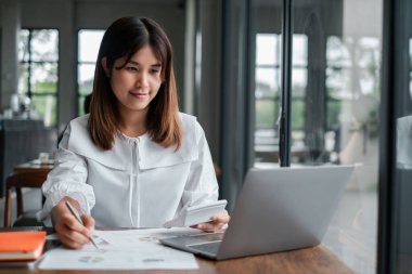 Beyaz bluzlu genç bir kadın dizüstü bilgisayar üzerinde çalışıyor ve modern ofis ortamında geniş pencereli belgeler hazırlıyor..