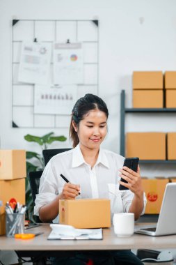 Çevrimiçi işlerini yürüten genç bir kadın, akıllı telefon kullanıyor ve paketleri modern bir ofis ortamında hazırlıyor..