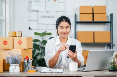 Akıllı telefon ve laptop kullanan genç bir kadın, karton kutularla çevrili, küçük bir işletme ya da e-ticaret ortamını işaret ediyor..