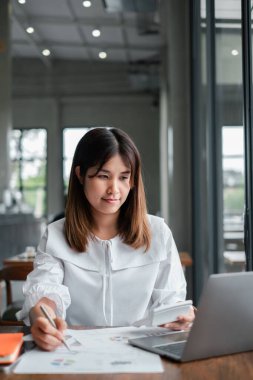 Modern ofis ortamında dizüstü bilgisayarı üzerinde çalışan genç profesyonel bir kadın doğal ışık ve geniş pencereli..