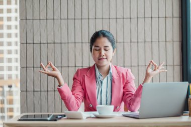 Pembe ceketli bir iş kadını modern bir ofiste masasında meditasyon yapıyor. Laptop ve kahve fincanı ile farkındalık ve stres giderme alıştırması yapıyor..