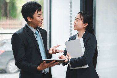 Modern ofis ortamında dijital tablet ve dokümanları elinde tutarken resmi giyinmiş iki iş adamı işi tartışıyor..