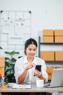 Beyaz gömlekli genç bir kadın ofis masasında dizüstü bilgisayarı, nakliye kutuları ve ofis malzemeleriyle akıllı telefon kullanıyor..