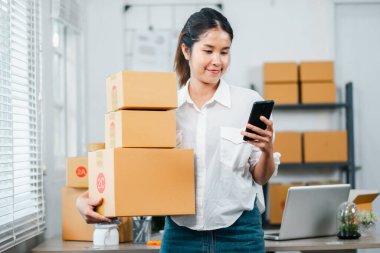İnternet üzerinden sipariş vermek için akıllı telefon kullanan genç bir kadın bir ofis ortamında kutu tutarken ve paketlerken.