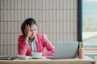 Pembe ceketli genç bir iş kadını ofis masasında dizüstü bilgisayarıyla çalışıyor. Yanında bir fincan kahve ve kırtasiye malzemesiyle düşünceli görünüyor..