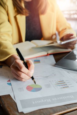 Kalem tutan bir iş kadınının el resmi. Ofiste dizüstü bilgisayar ve defterle mali raporları analiz ediyor..