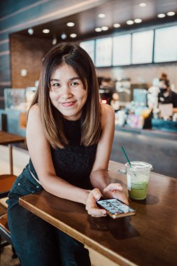 Genç bir kadın modern bir kahve dükkanında masada oturuyor, akıllı telefonunu kullanarak gülümsüyor ve yeşil buzlu içkinin tadını çıkarıyor..