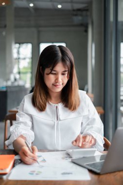 Beyaz bluzlu genç bir kadın modern bir ofis masasında finansal belgeler üzerinde çalışıyor. Laptop ve hesap makinesi kullanıyor..
