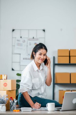 Evden çalışan genç bir kadın, bir dizüstü bilgisayar ve telefonla birden fazla iş yapıyor, etrafı kargo kutularıyla çevrili, küçük işletmeleri ve e-ticareti temsil ediyor..