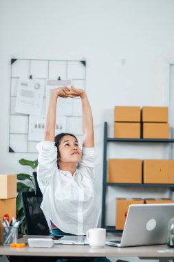 Genç bir kadın, çalışma saatlerinin yoğun olduğunu gösteren mukavva kutular ve dizüstü bilgisayarlarla çevrili modern ofis ortamında masasına uzanır..