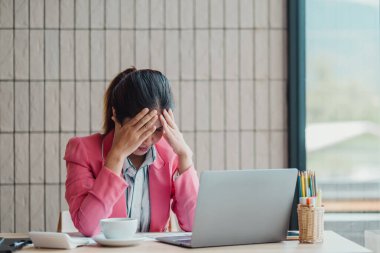 Pembe ceketli bir iş kadını, elinde kahve fincanı ve kırtasiyesiyle ofis masasında dizüstü bilgisayarıyla çalışırken stresli görünür..