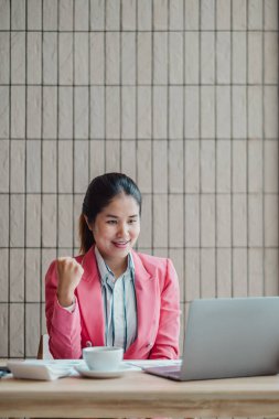 Pembe ceketli genç profesyonel bir kadın, modern ofis ortamında dizüstü bilgisayarı üzerinde çalışırken başarısını kutluyor..