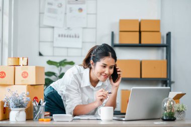 Küçük işletmesini evden idare eden genç bir kadın, bir telefon görüşmesi ve dizüstü bilgisayarla birden fazla iş yapıyor, paket kutularıyla çevrili..