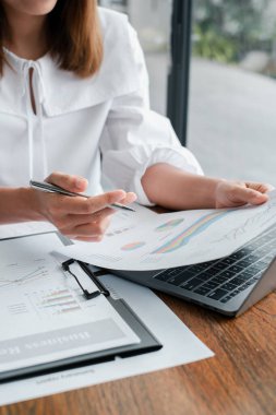 Çağdaş bir ofis ortamında, kağıt ve dizüstü bilgisayardaki mali grafikleri ve grafikleri inceleyen bir iş kadınının yakın çekimi.