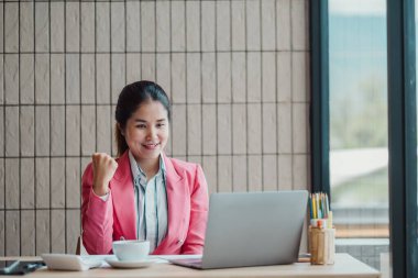 Pembe ceketli, modern bir ofiste dizüstü bilgisayarda çalışan kendine güvenen genç bir kadın başarı ve memnuniyeti ifade ediyor..