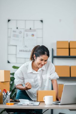 Modern bir ofiste dizüstü bilgisayarla çalışan beyaz gömlekli genç bir iş kadını. Etrafında karton kutular ve ofis malzemeleri var..