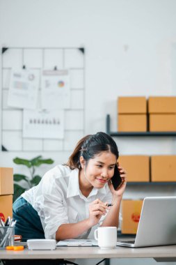 Ofisinde birden fazla iş yapan genç bir kadın, telefonla konuşuyor ve dizüstü bilgisayarla çalışıyor, etrafında posta kutuları ve ofis malzemeleri var..
