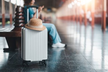 Bir gezgin, beyaz bir bavul ve hasır şapkayla tren istasyonunda oturuyor, akıllı telefon kullanıyor, sıcak güneş ışığında banyo yapıyor..