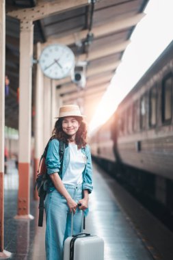 Bavullu genç bir kadın gezgin tren istasyonunda duruyor, gün doğumunda gülümseyerek ve yolculuğuna hazır olarak..