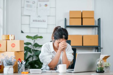 Genç bir kadın çalışma masasında dizüstü bilgisayarı ve modern ofis ortamında kutularla otururken stresli görünüyor..