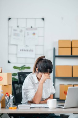 Genç bir iş kadını, masasında dizüstü bilgisayarla çalışırken ve modern bir ofis ortamında kutu taşırken stresli hissediyor..