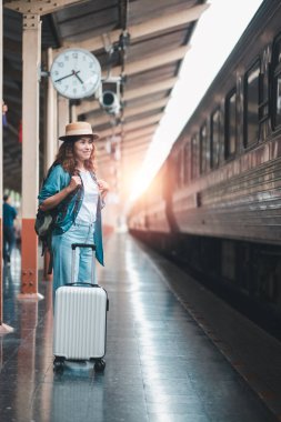 Sırt çantası ve bavulu olan genç bir kadın gün doğarken tren istasyonunda bekliyor. Yolculuk macerasına hazır..