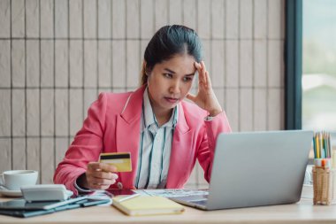 Pembe ceketli bir kadın, elinde kredi kartıyla ofis ortamında dizüstü bilgisayarına bakarken stresli görünür..
